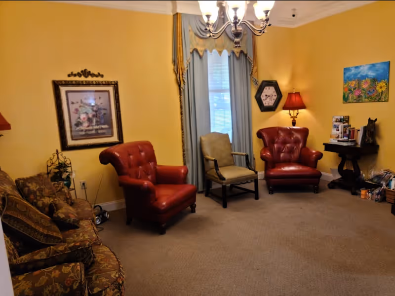 Cozy sitting room with two red leather armchairs, a patterned sofa, a green chair by a curtained window, wall art and side tables.