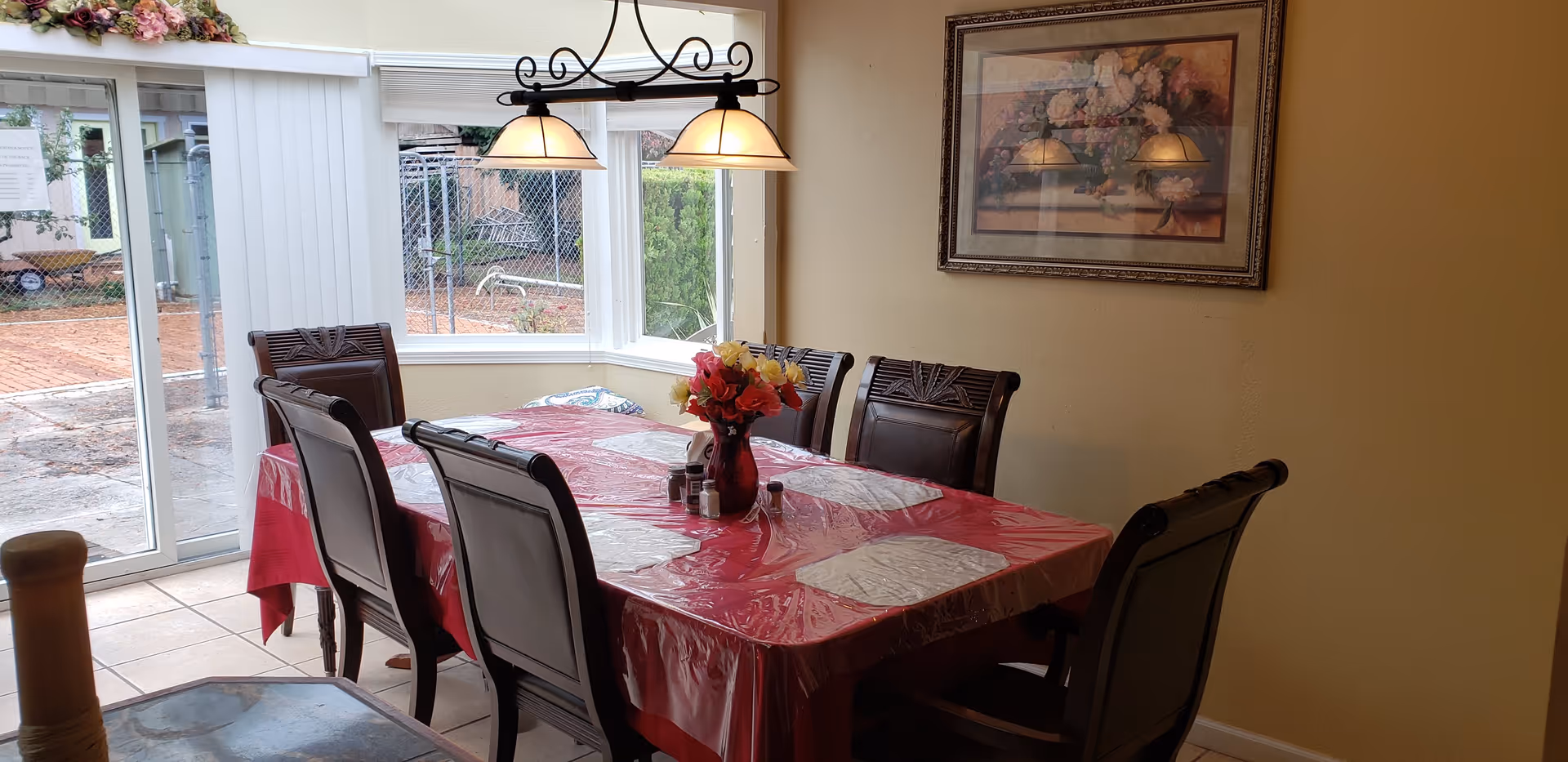 Dining room with a rectangular table covered with a red plastic tablecloth and six wooden chairs around it. A vase with colorful flowers and salt and pepper shakers are on the table. There is a hanging light fixture with two lamps above the table. Large windows and a sliding glass door provide a view of an outdoor area with a brick patio and some greenery. A framed floral painting hangs on the wall.