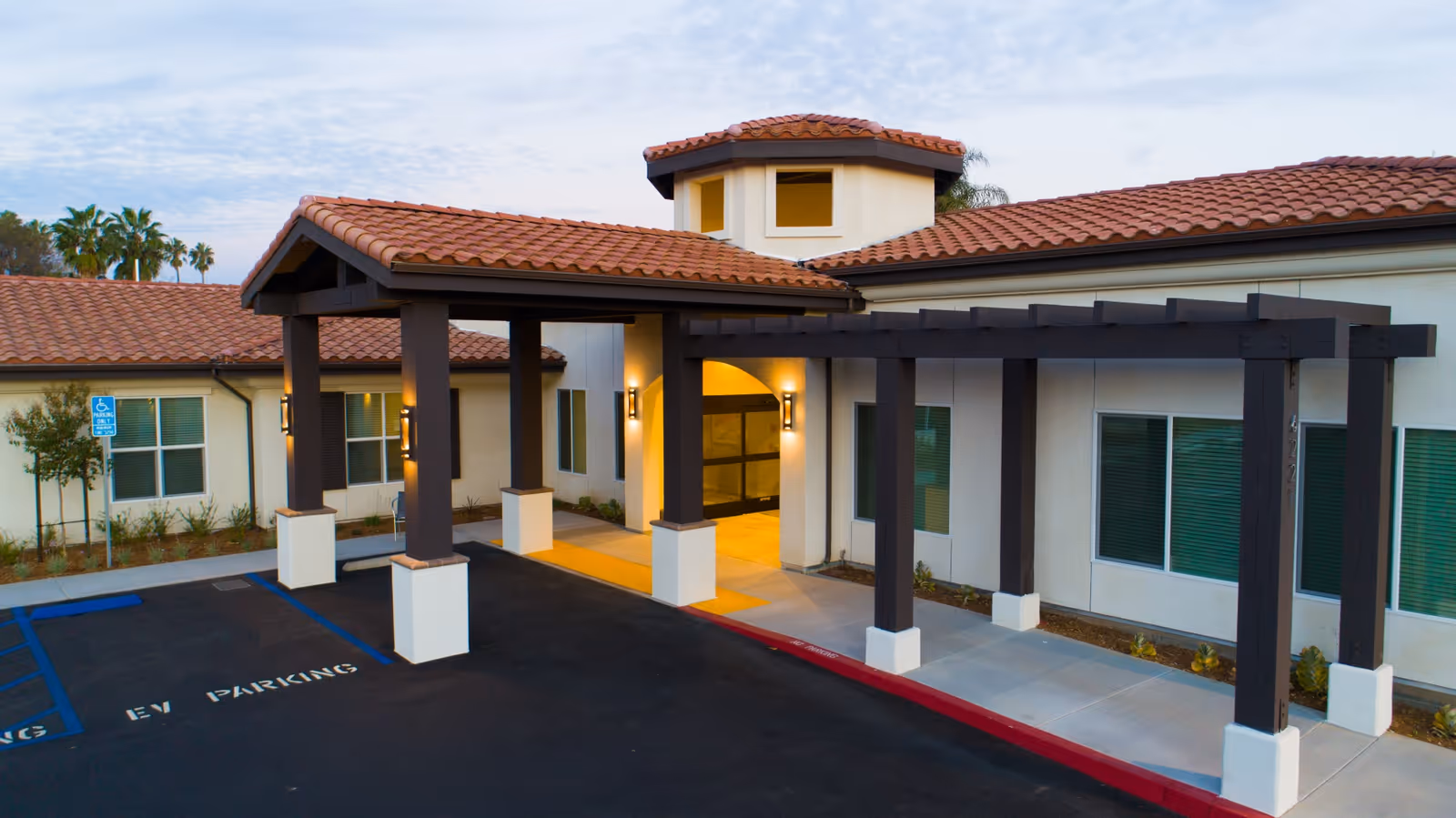 Entrance of The Preserve at Woodland Hills Assisted Living & Memory Care facility with a covered drop-off area, beige walls, red tile roof, and adjacent parking spaces including EV parking and a handicapped spot.