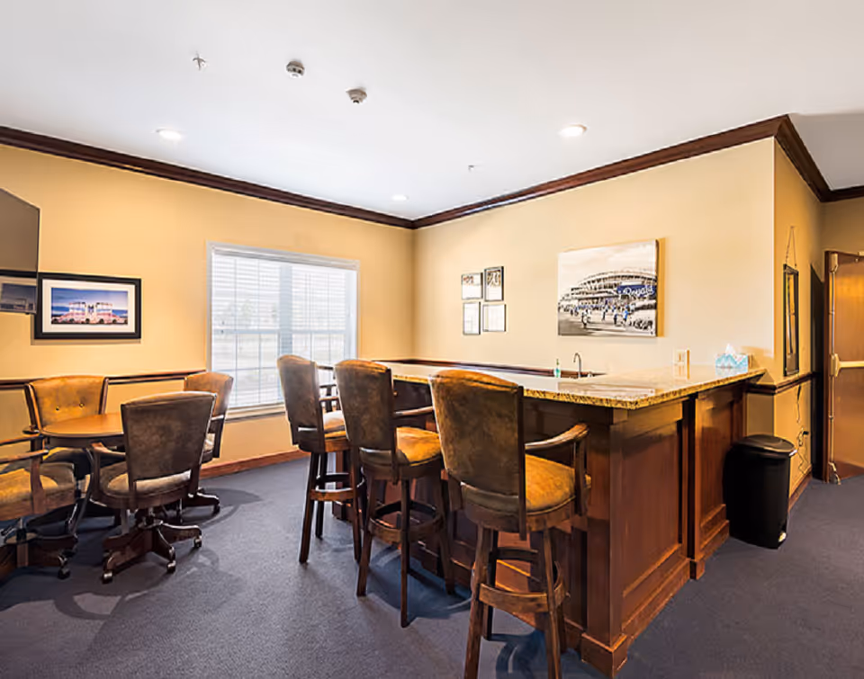 Interior common area featuring a wooden bar with high stools, a round table with chairs, and framed artwork on warm-toned walls.