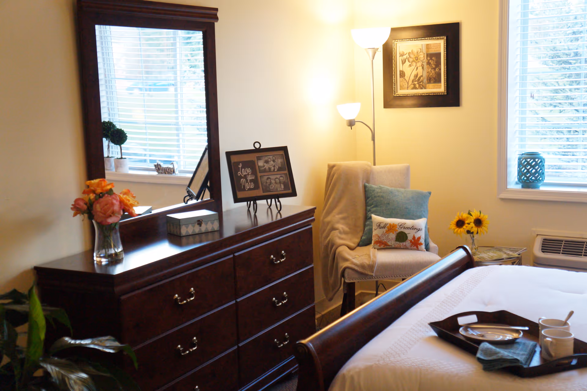 A cozy bedroom in a senior living facility featuring a wooden dresser with a large mirror, a vase with flowers, framed photos, and decorative items. Next to the dresser is a comfortable chair with pillows and a throw blanket, a floor lamp, and a framed floral picture on the wall. The bed has a tray with cups and a plate, and there are windows with blinds letting in natural light.