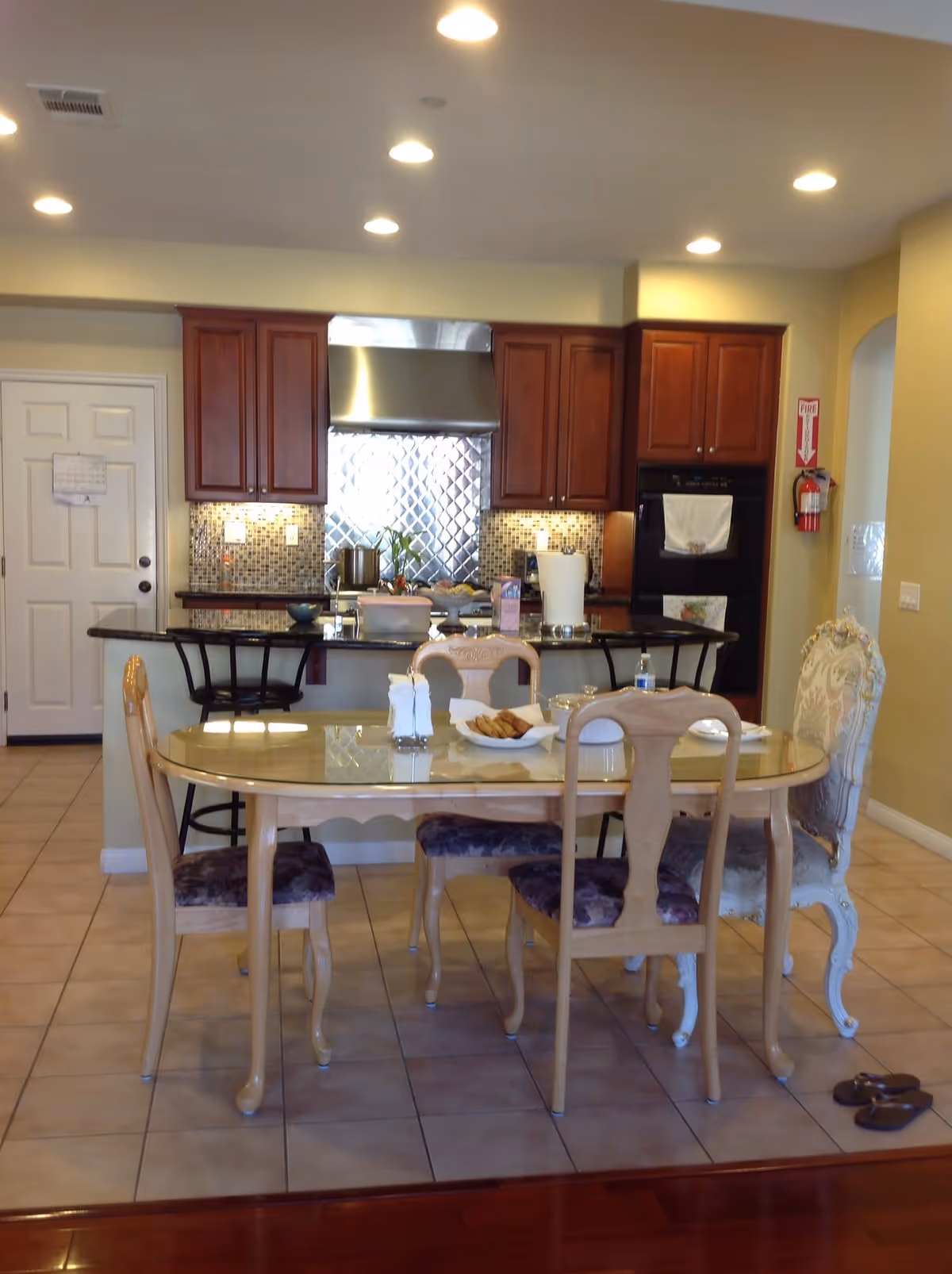 Interior view of a kitchen and dining area in a senior living facility. The kitchen features wooden cabinets, a stainless steel range hood, a tiled backsplash, and a black countertop with bar stools. In front of the kitchen is a glass-top dining table with four chairs, one of which is upholstered with a floral pattern. On the table, there are plates, a bowl of cookies, a water bottle, and napkins. The floor is tiled, and the walls are painted light yellow. A fire extinguisher is mounted on the wall near the kitchen.