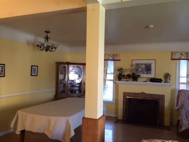 Interior view of a senior living facility room with yellow walls. The room features a dining table covered with a beige tablecloth, a wooden china cabinet with glass doors, a fireplace with decorative items on the mantel, two windows with floral valances, and a chandelier hanging from the ceiling.