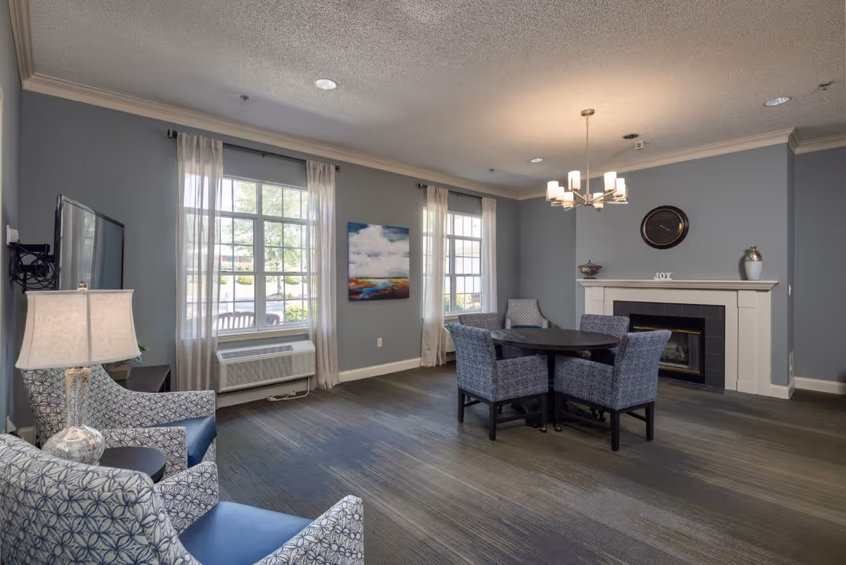 A cozy living room area with two patterned armchairs and a small table with a lamp on the left, a round dining table with four upholstered chairs in the center, and a fireplace with a clock and decorative items on the mantel. The room has two large windows with sheer curtains letting in natural light, blue-gray walls, and a modern chandelier hanging from the ceiling.