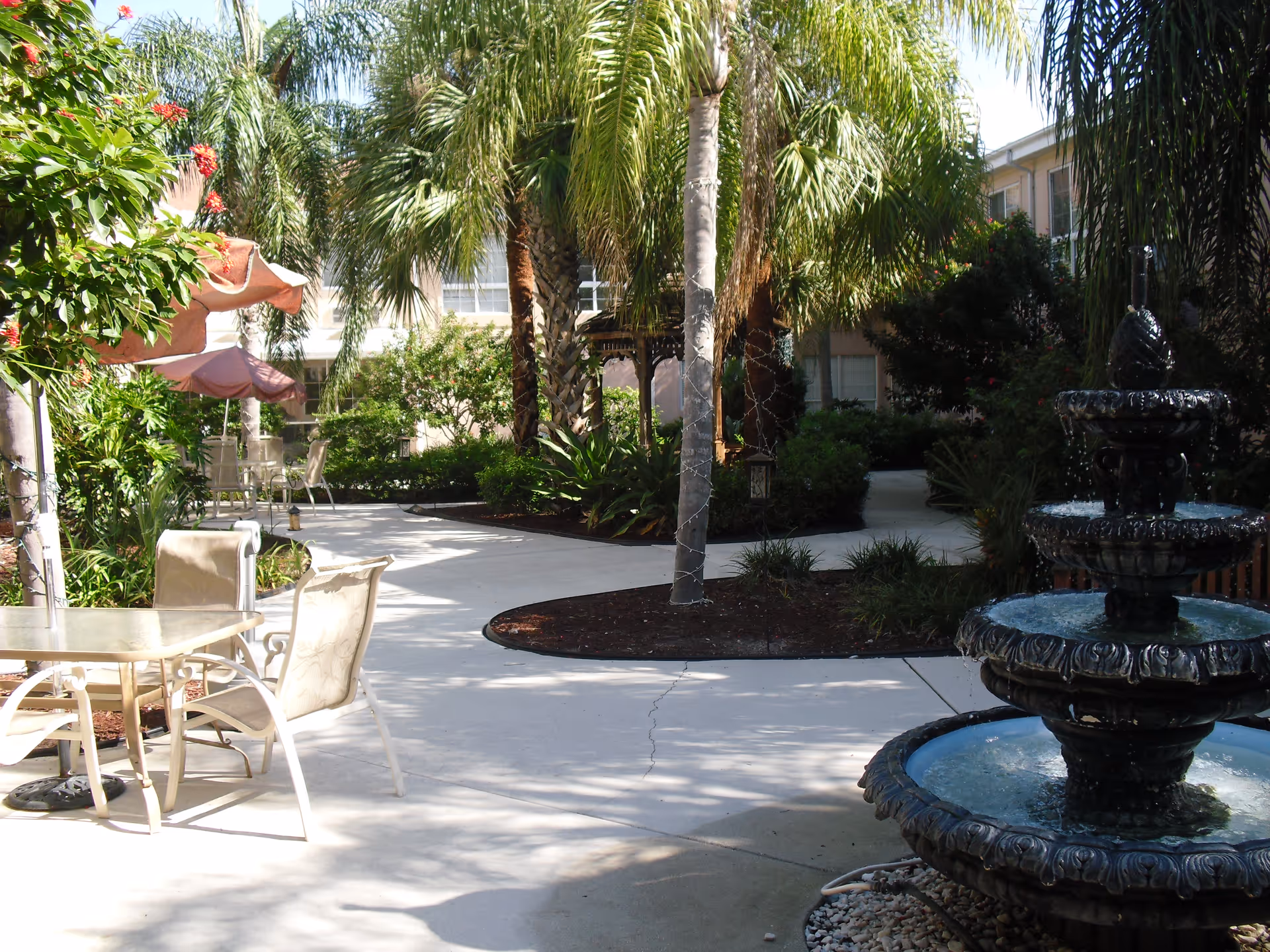 A sunny courtyard with palm trees, patio tables and umbrellas, and a tiered fountain near a building.