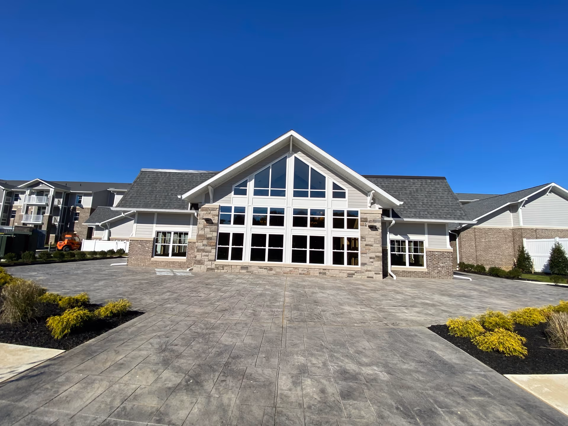 Exterior view of a modern retirement community building with large windows and a peaked roof under a clear blue sky. The paved area in front is spacious with landscaped greenery on the sides.