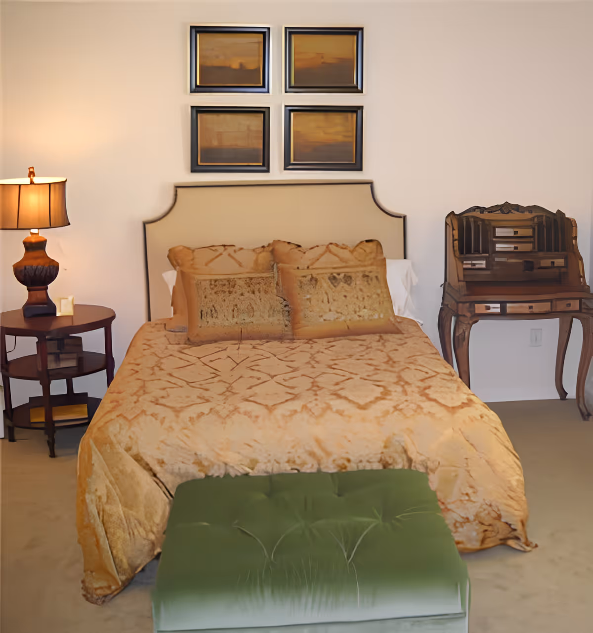A bedroom with a beige upholstered headboard, a bed covered with a patterned beige bedspread and matching pillows, a green tufted bench at the foot of the bed, a wooden side table with a lamp on the left, and a vintage wooden writing desk on the right. Four framed abstract paintings hang on the wall above the headboard.