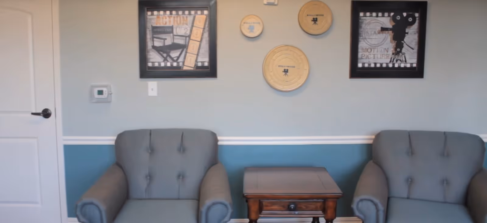 Two gray upholstered armchairs with button tufting are positioned on either side of a small wooden side table with a drawer. The wall behind them is painted light gray on the upper half and blue on the lower half, separated by white trim. On the wall are two framed pictures featuring film-related themes and three round wooden plaques with the word 'Hollywood' and film icons. A white door with a black handle and a thermostat are visible on the left side of the image.
