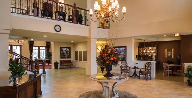 Spacious and elegant senior living facility lobby with a round table holding a large floral arrangement in the center. The area features tiled floors, a chandelier, comfortable seating areas, artwork on the walls, and a staircase leading to an upper balcony with additional seating.