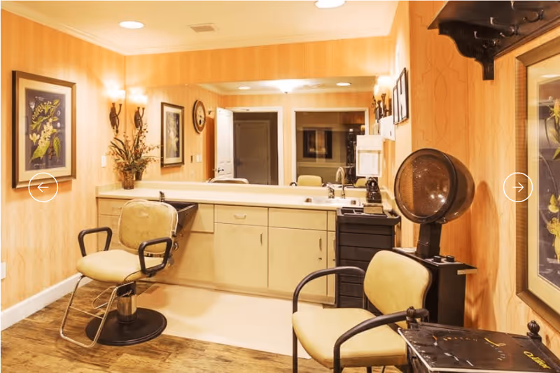Interior of a salon area in an assisted living facility with two vintage-style salon chairs, a hair dryer, a large mirror, framed floral artwork on the walls, and warm yellow lighting.