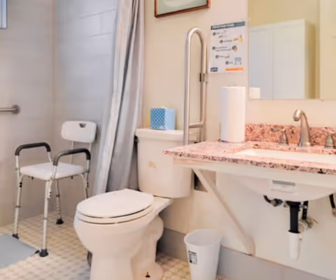 A bathroom designed for accessibility featuring a toilet with a grab bar, a shower area with a shower chair, a sink with a granite countertop, a paper towel roll, a tissue box, and a mirror above the sink.