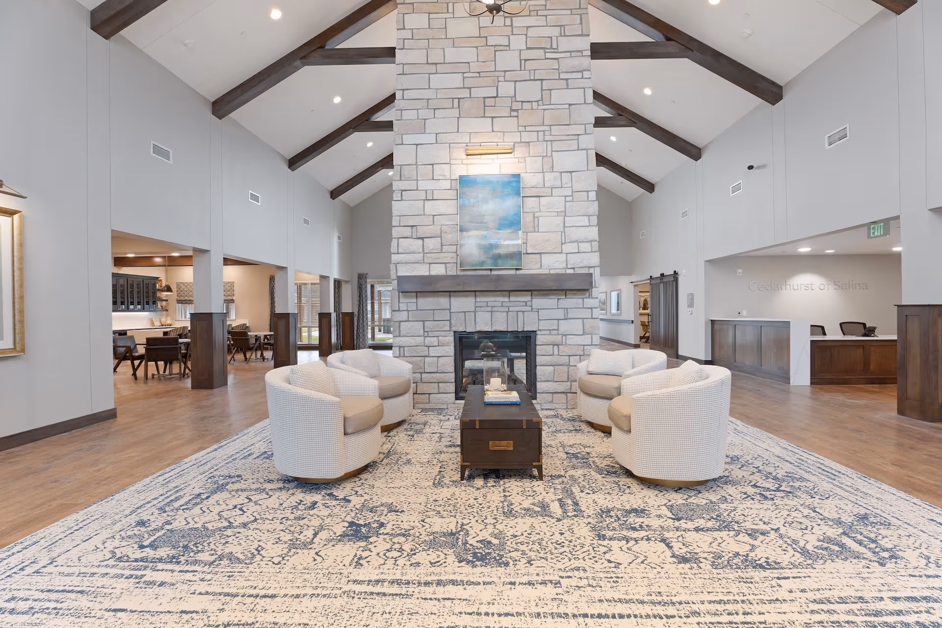 Spacious senior living lounge with a central stone fireplace, four round armchairs around a coffee table on a patterned rug and vaulted ceiling with exposed beams.