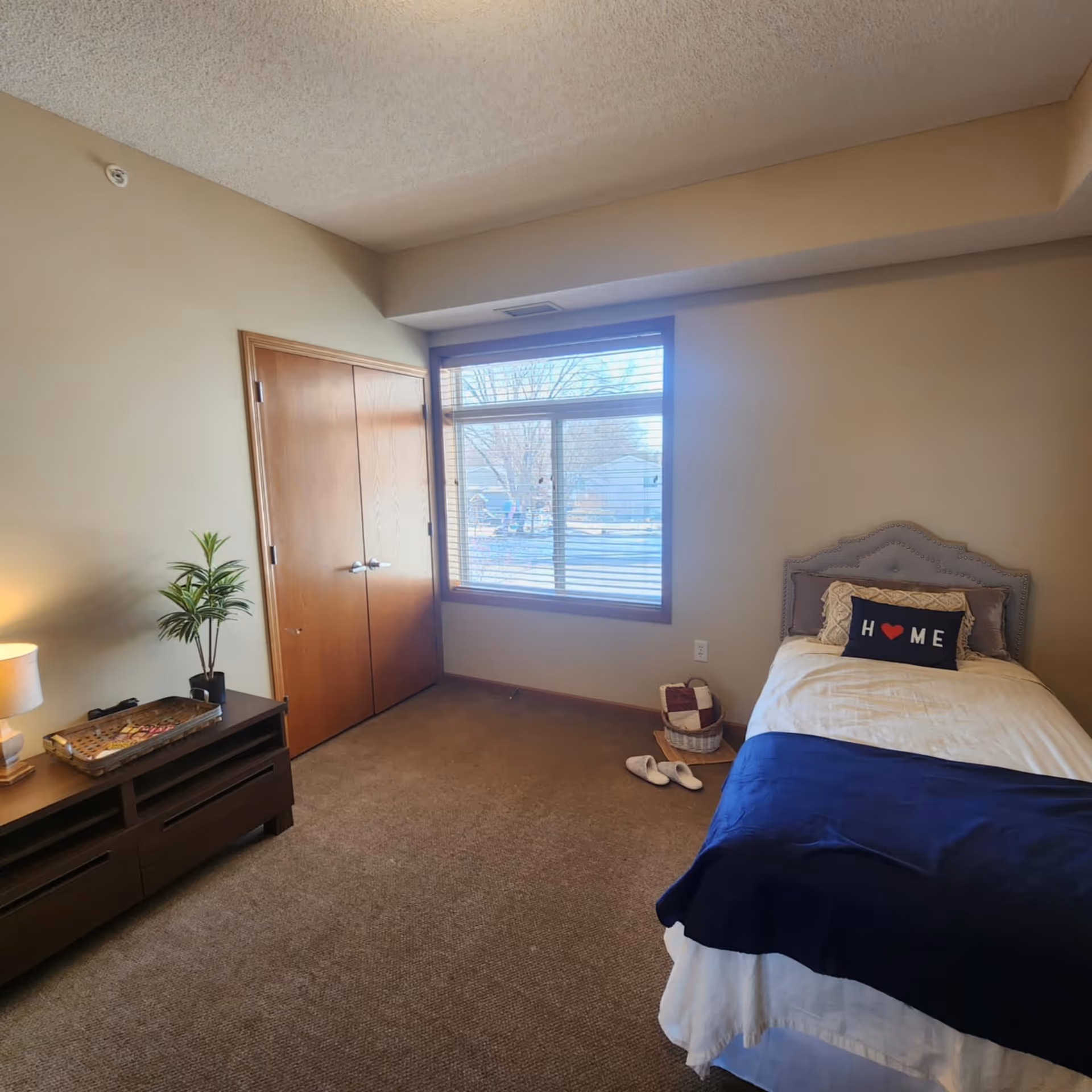 A cozy bedroom with a single bed featuring a gray upholstered headboard, white bedding, and a navy blue throw blanket. A decorative pillow on the bed reads 'HOME' with a red heart symbol. Next to the bed is a basket with a folded blanket and a pair of white slippers on the carpeted floor. A wooden dresser with a lamp and a small potted plant is positioned against the opposite wall. There is a large window with blinds letting in natural light and a wooden double-door closet beside it.