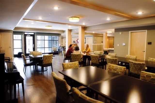 A spacious dining room with multiple dark wooden tables and cushioned chairs arranged neatly. Several people are seated at one of the tables engaged in conversation. The room has large windows allowing natural light to enter, and the floor is wooden. The ceiling has recessed lighting and modern light fixtures.