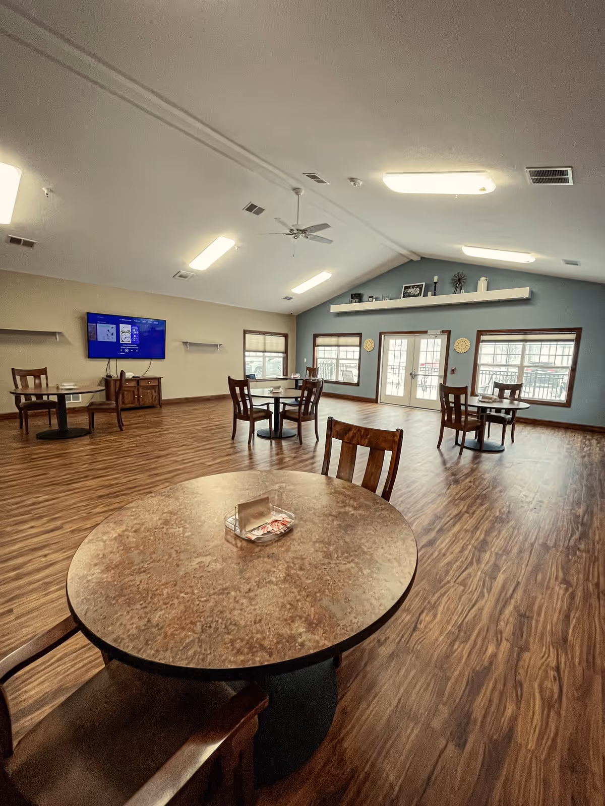 A spacious room with several round tables and wooden chairs arranged on a wood-patterned floor. The room has a high ceiling with ceiling fans and fluorescent lights. There are multiple windows and a set of double glass doors on one wall, allowing natural light to enter. A flat-screen TV is mounted on one wall above a small wooden cabinet. The walls are painted in neutral tones with one accent wall in blue, decorated with a shelf holding various decorative items.