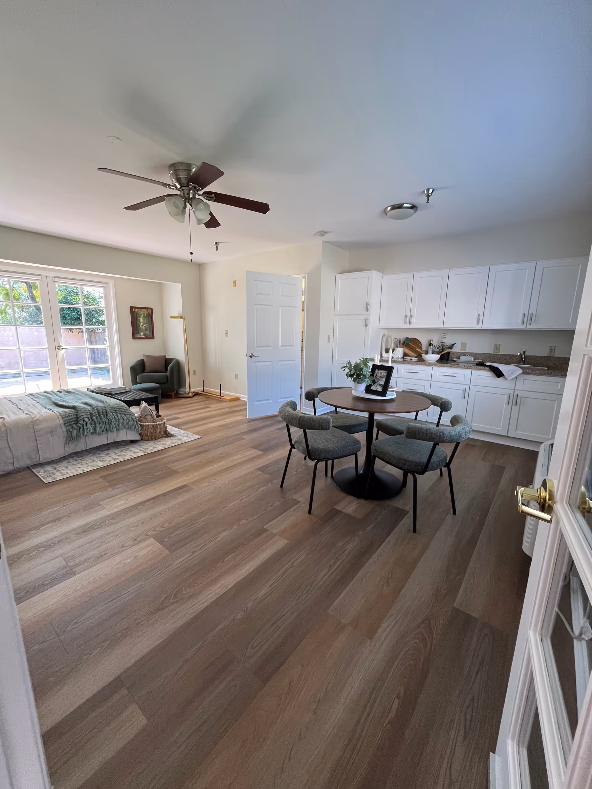A bright, spacious studio apartment with a bed on the left side near large windows, a small round dining table with four chairs in the center, and a kitchenette with white cabinets and a countertop on the right. There is a ceiling fan with lights above, a green armchair in the corner near the windows, and wooden flooring throughout.