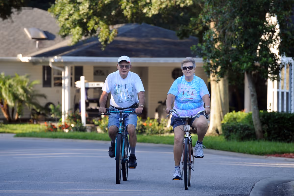 An elderly man and woman riding bicycles on a paved road in a residential area with houses and greenery in the background. Both are wearing casual clothes and sunglasses.