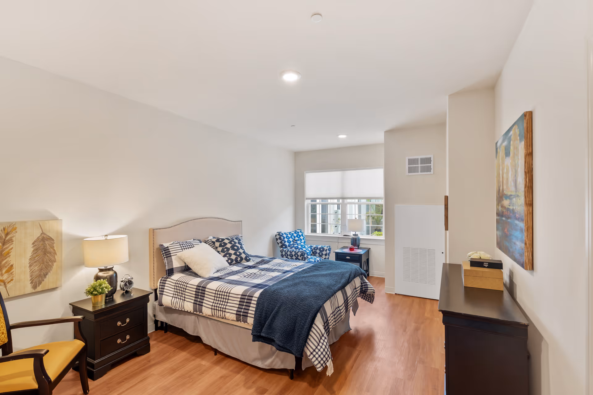 A well-lit bedroom with a bed dressed in blue and white checkered bedding and a dark blue throw blanket. There is a nightstand with a lamp, a small plant, and a clock beside the bed. A patterned armchair is positioned near a window with white blinds. The room has wooden flooring, a dresser with decorative boxes, and a painting on the wall.