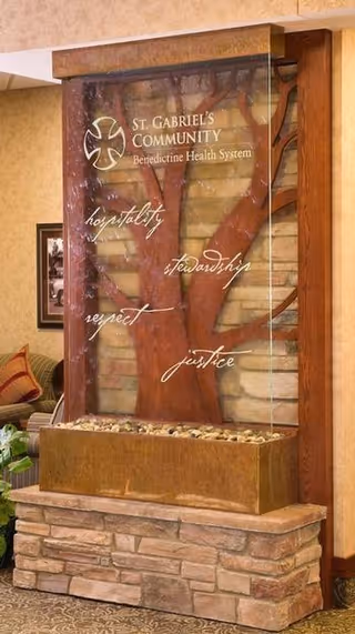 Indoor decorative water feature with a tree design and inscriptions including 'St. Gabriel's Community Benedictine Health System' and words like hospitality, stewardship, respect, and justice. The feature is set against a stone base with a seating area and framed picture visible in the background.