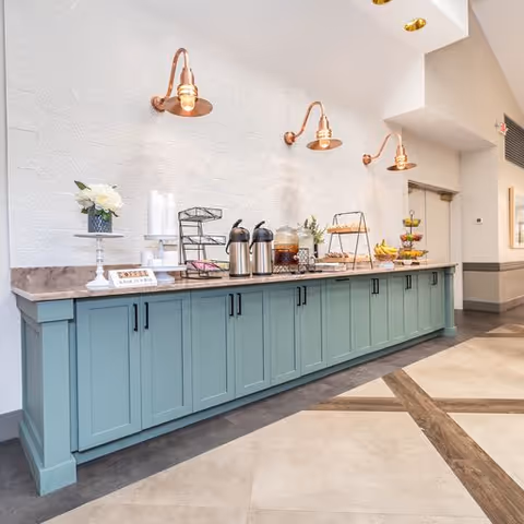 A refreshment area with a long teal cabinet topped with a marble countertop. On the counter are coffee dispensers, a glass container with iced tea, a tiered tray with bananas and apples, a small flower arrangement, and a sign that says 'Coffee is always a good idea.' The wall behind has a white textured surface with three copper wall-mounted light fixtures. The floor has a geometric pattern with light and dark tiles.