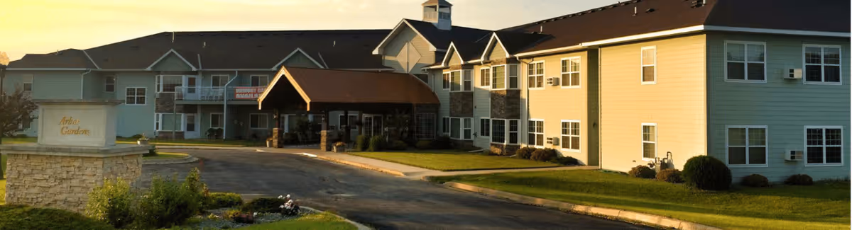 Exterior view of Arbor Garden Place, a two-story senior living facility with beige siding and multiple windows. The building has a covered entrance with a brown roof and a driveway leading up to it. There is a stone sign in the foreground with the name Arbor Garden, surrounded by greenery and landscaping.
