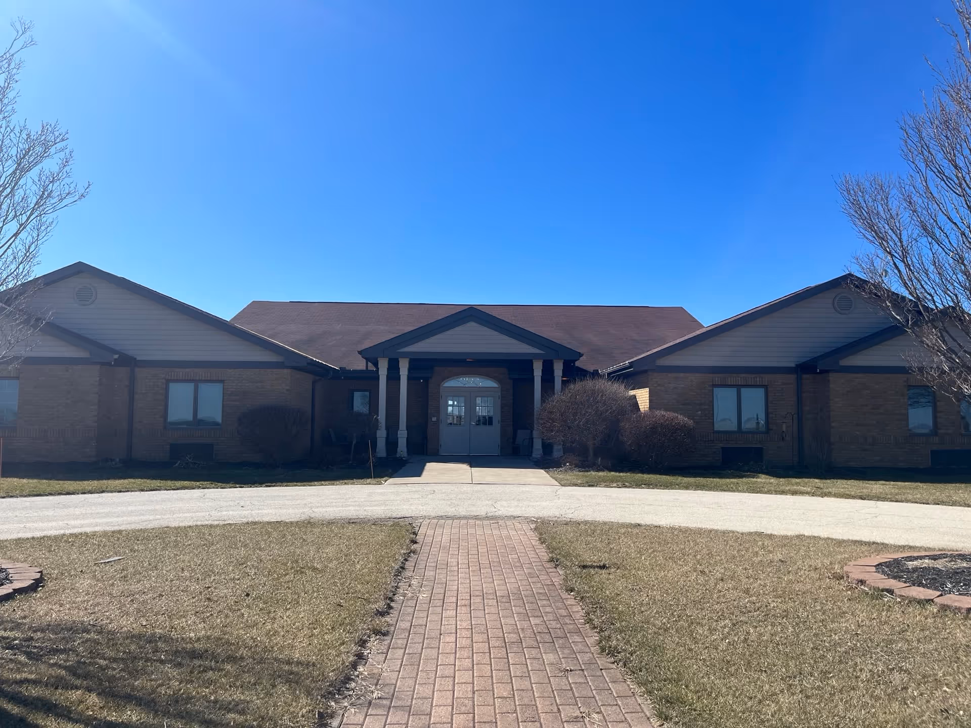 Front exterior view of a single-story brick building with a peaked roof and a central entrance featuring double doors and white columns. A brick pathway leads up to the entrance, flanked by grass and leafless trees under a clear blue sky.