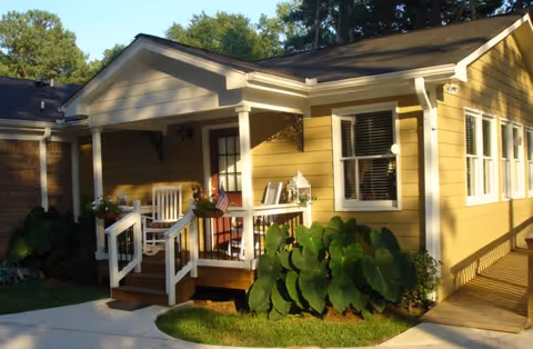 Front porch of a yellow single-story building with white railings, rocking chairs, potted plants, and large green foliage.