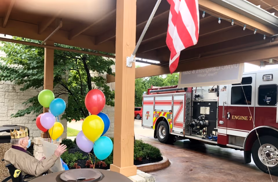 A senior in a wheelchair wearing a paper crown and mask watches a fire engine arrive under a covered entrance decorated with colorful balloons and an American flag.
