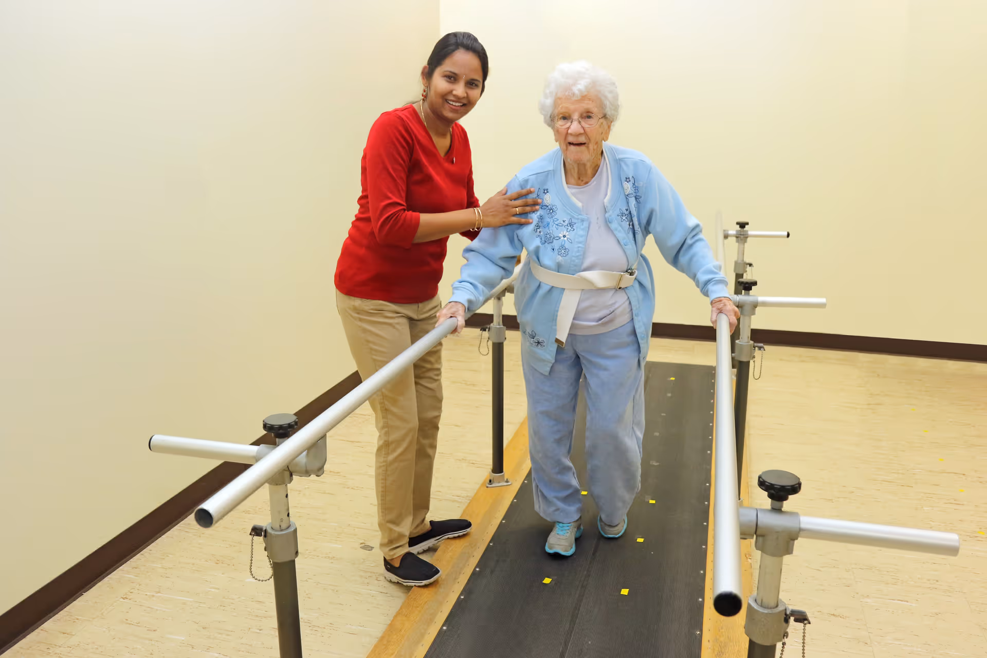 An elderly woman wearing a light blue jacket and pants is walking with the support of parallel bars in a rehabilitation or physical therapy setting. A younger woman in a red sweater and beige pants is standing beside her, smiling and providing assistance.