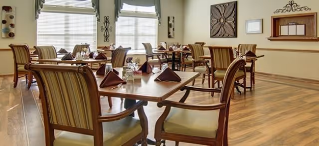 Dining room with multiple tables set for meals, wooden chairs, folded napkins, glassware, and large windows.