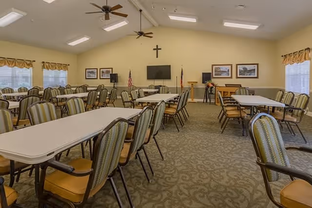 A large meeting or dining room with multiple rectangular tables and green cushioned chairs arranged neatly. The room has beige walls, carpeted floor, ceiling fans, and several windows with valances. At the front of the room, there is a wall-mounted TV, a cross, an American flag, a Texas state flag, framed pictures, a podium, and a piano.