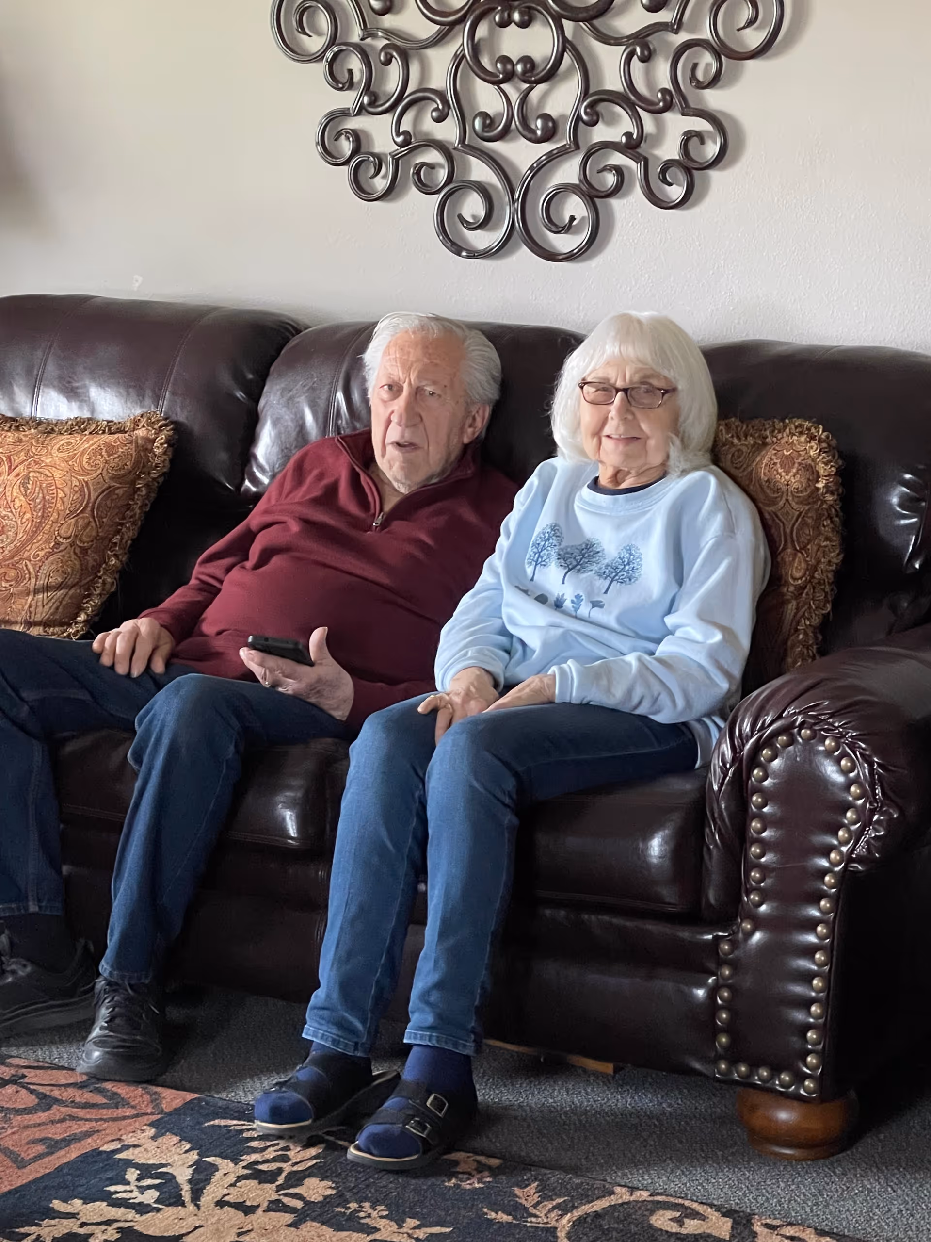 An elderly man and woman sitting together on a dark brown leather couch in a living room. The man is wearing a maroon sweater and dark pants, holding a remote control, while the woman is wearing glasses, a light blue sweatshirt with a tree design, and blue jeans. Behind them on the wall is a decorative metal wall art piece, and there are patterned cushions on the couch.