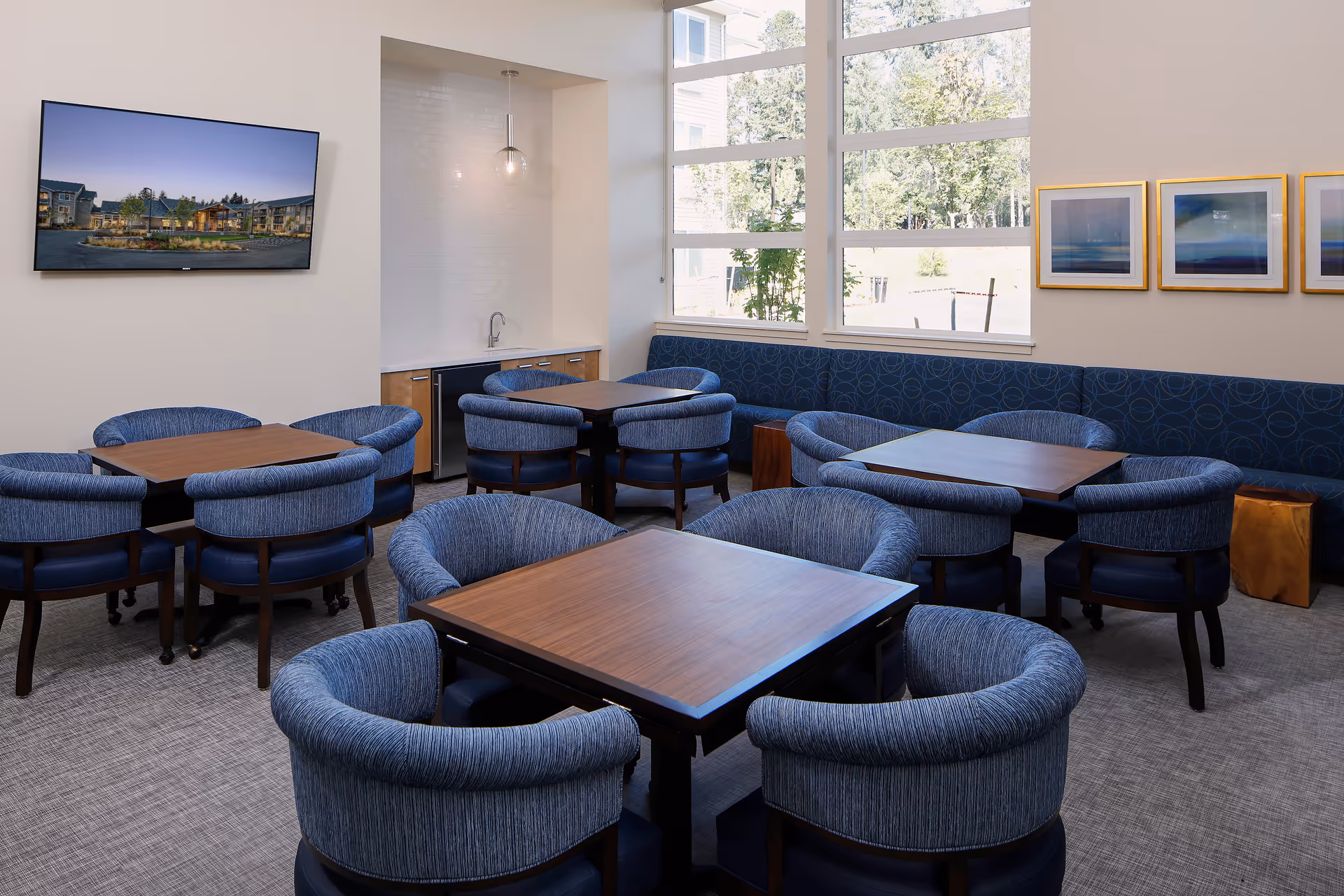 Bright communal dining area with several square wooden tables and blue upholstered chairs, a wall-mounted TV, and large windows.