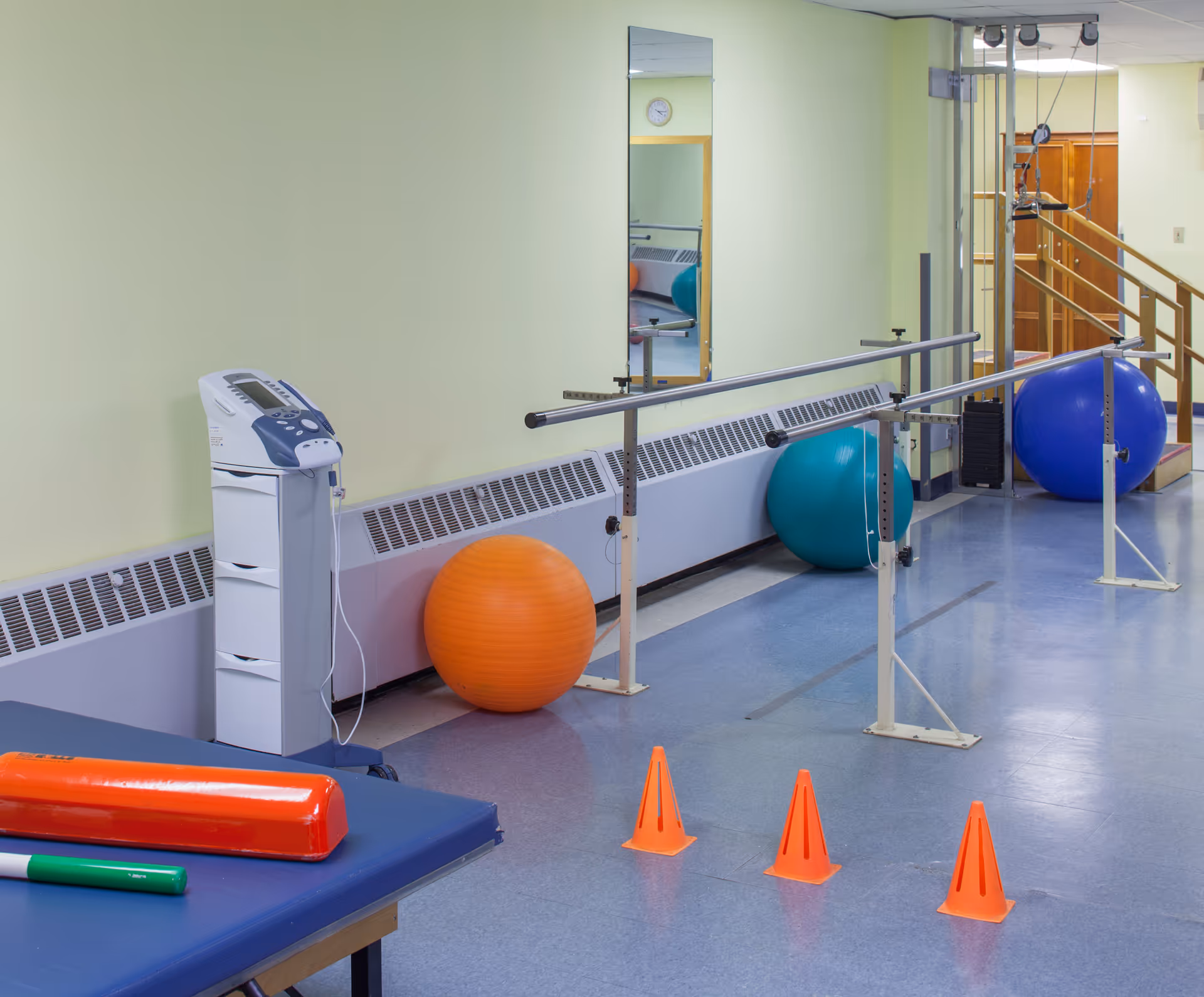 Physical therapy room with parallel bars for walking exercises, exercise balls in orange, teal, and blue, orange cones on the floor, a therapy table with foam rollers, and a medical device against a light green wall with a mirror.