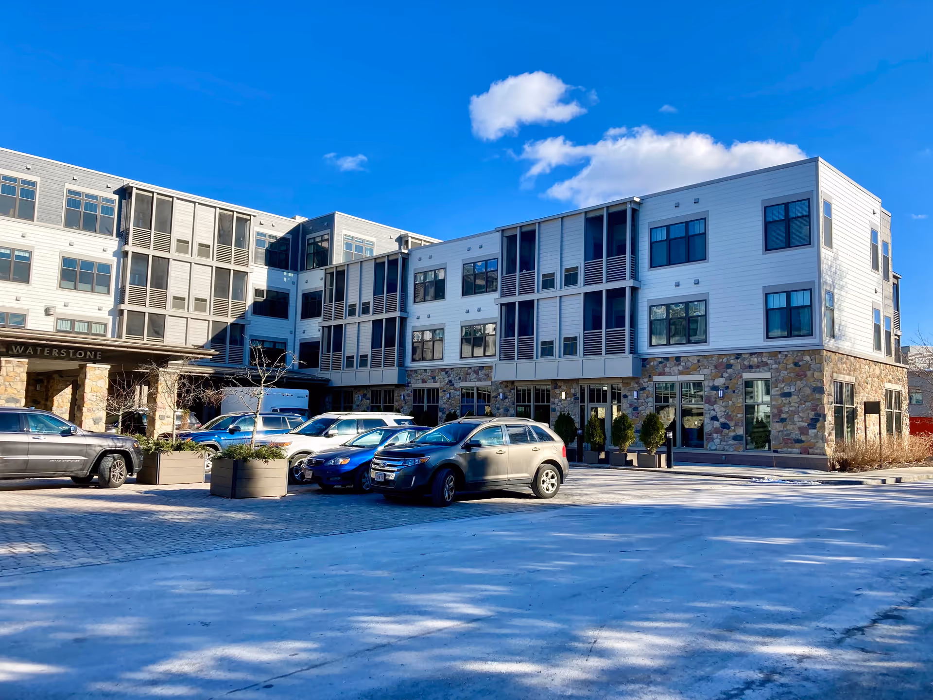 Exterior view of a modern three-story senior living facility named Waterstone At Wellesley with a stone and siding facade, multiple windows, and a parking area with several cars in front under a clear blue sky.