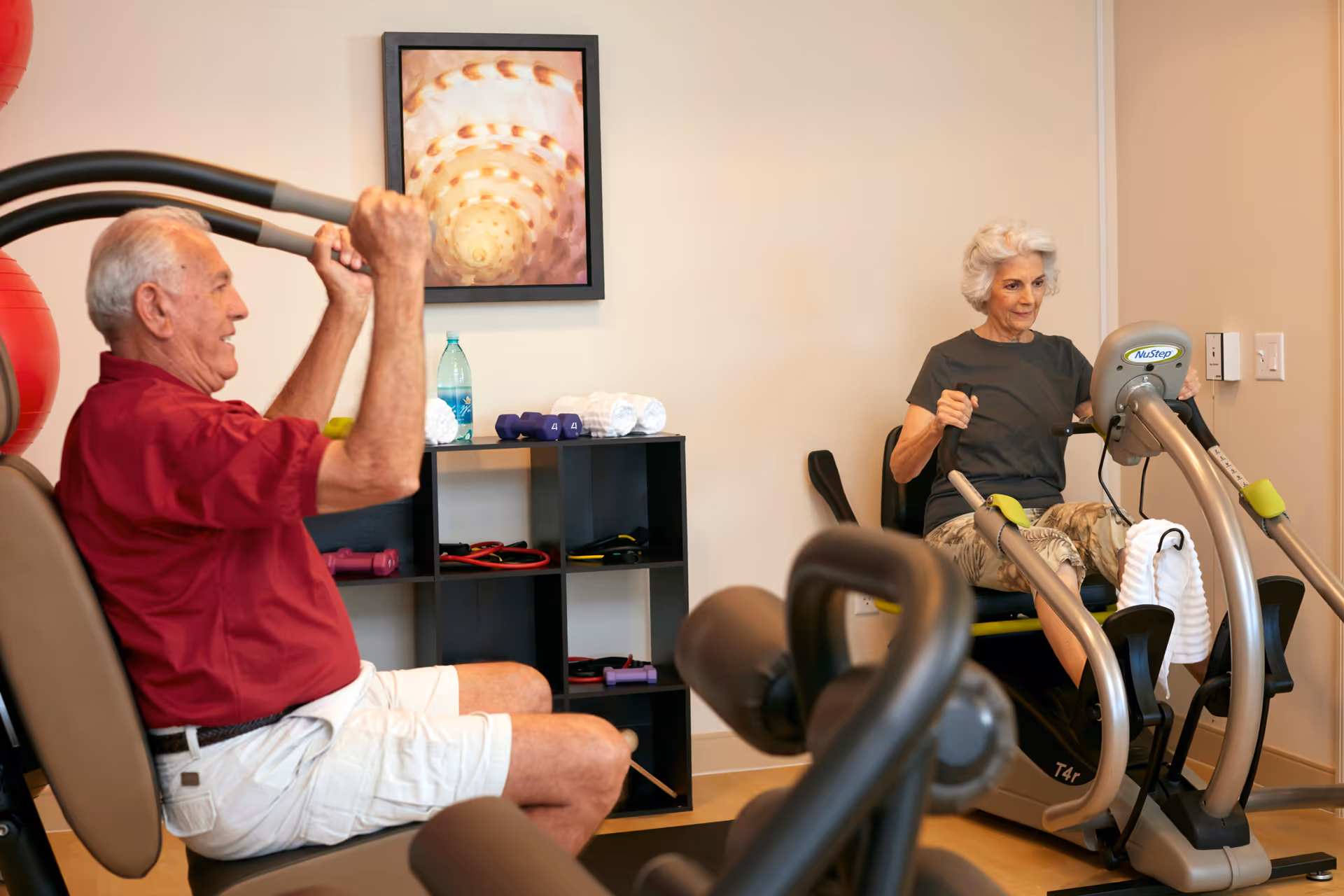 An elderly man and woman exercising on fitness machines in a well-lit room. The man is using a seated chest press machine, wearing a red shirt and white shorts. The woman is using a NuStep recumbent cross trainer, wearing a dark shirt and patterned pants. Behind them is a black shelving unit with small weights, towels, and a water bottle. A framed abstract artwork hangs on the wall.
