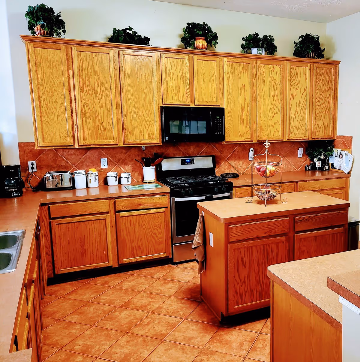 A kitchen with wooden cabinets and drawers, a stainless steel stove and microwave, a small kitchen island with a two-tiered fruit basket, and several potted plants on top of the upper cabinets. The floor is tiled with reddish-brown tiles, and there are various kitchen items like a toaster, coffee maker, and canisters on the countertops.
