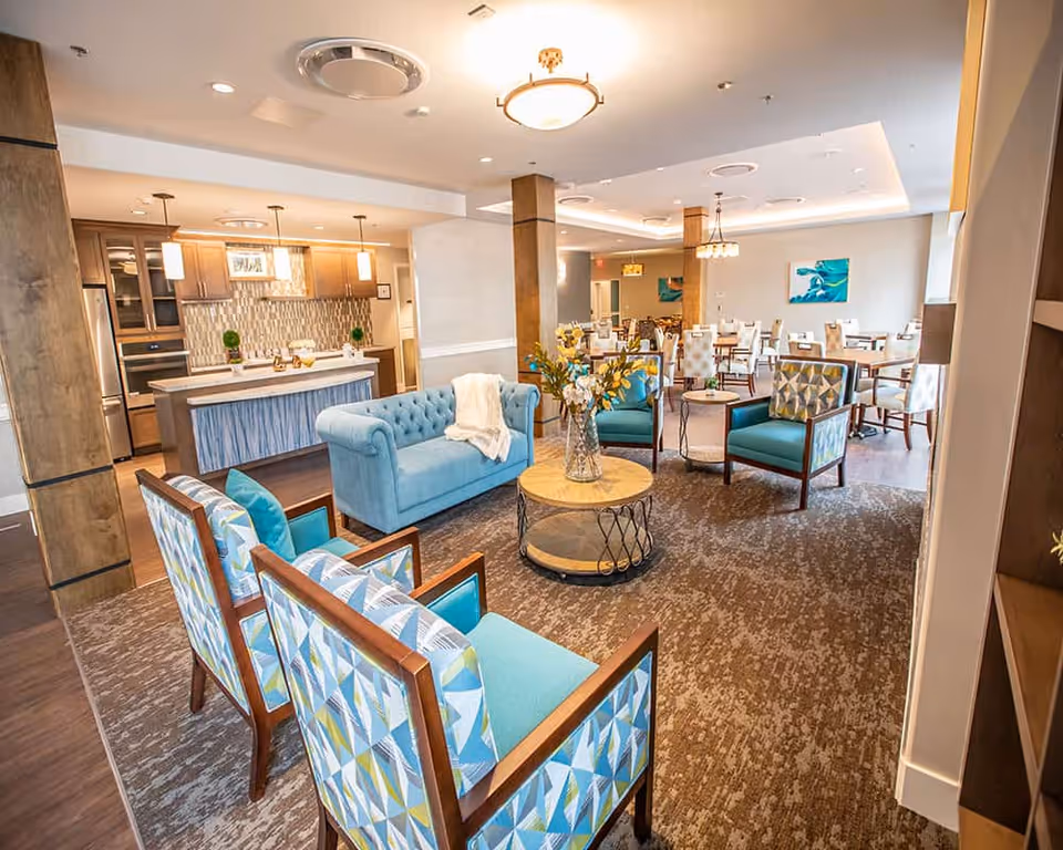 A bright and spacious common area in a senior living facility featuring a seating area with a blue sofa, patterned armchairs, a round wooden coffee table with a vase of flowers, and a dining area with multiple tables and chairs. The background includes a kitchen area with modern appliances and pendant lighting.