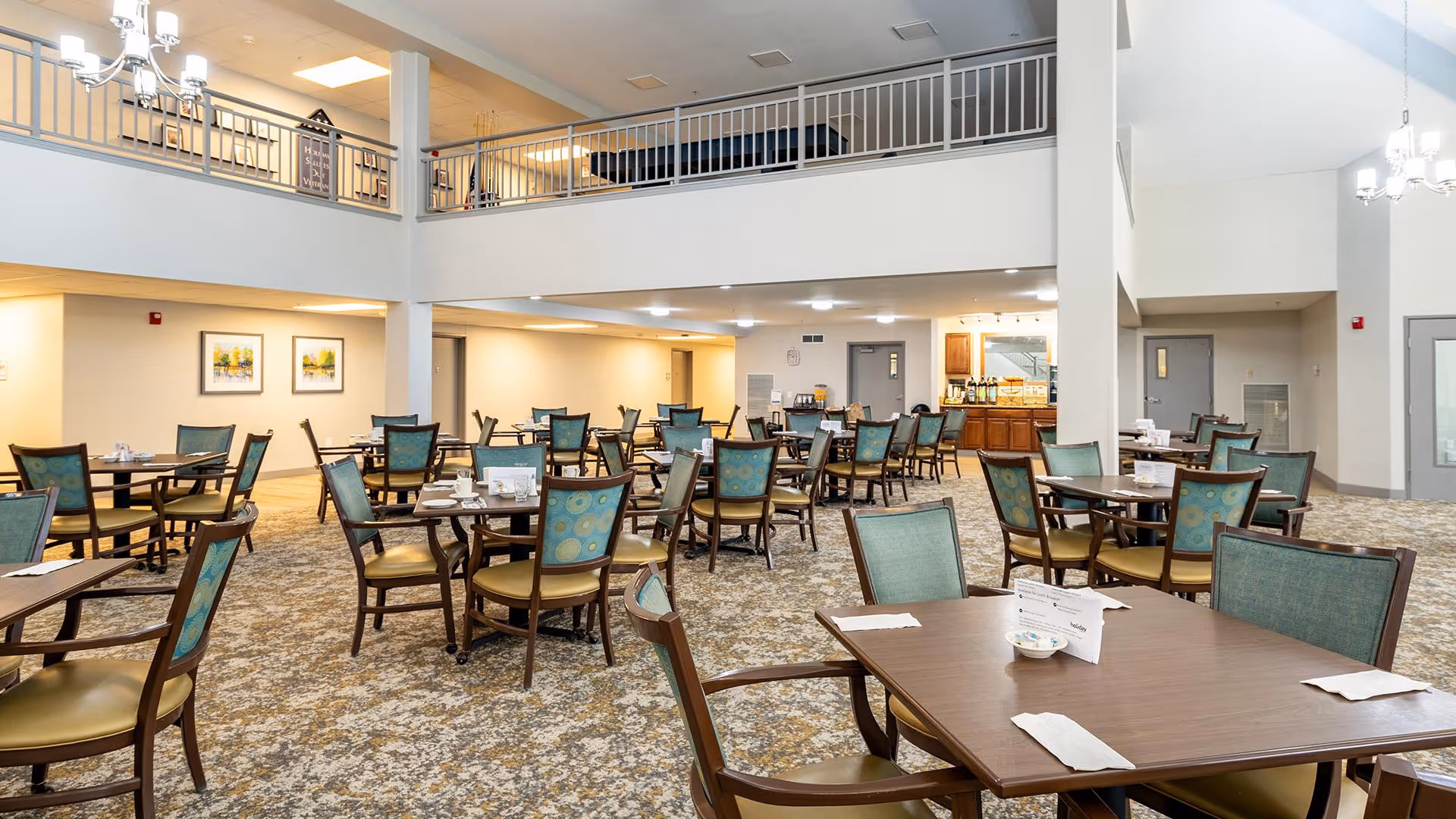 A spacious dining room with multiple wooden tables and chairs arranged neatly. The chairs have green and blue patterned upholstery. The room has a high ceiling with a second-floor balcony overlooking the area. There are chandeliers hanging from the ceiling and framed artwork on the walls. In the background, there is a counter area with coffee and beverage dispensers.