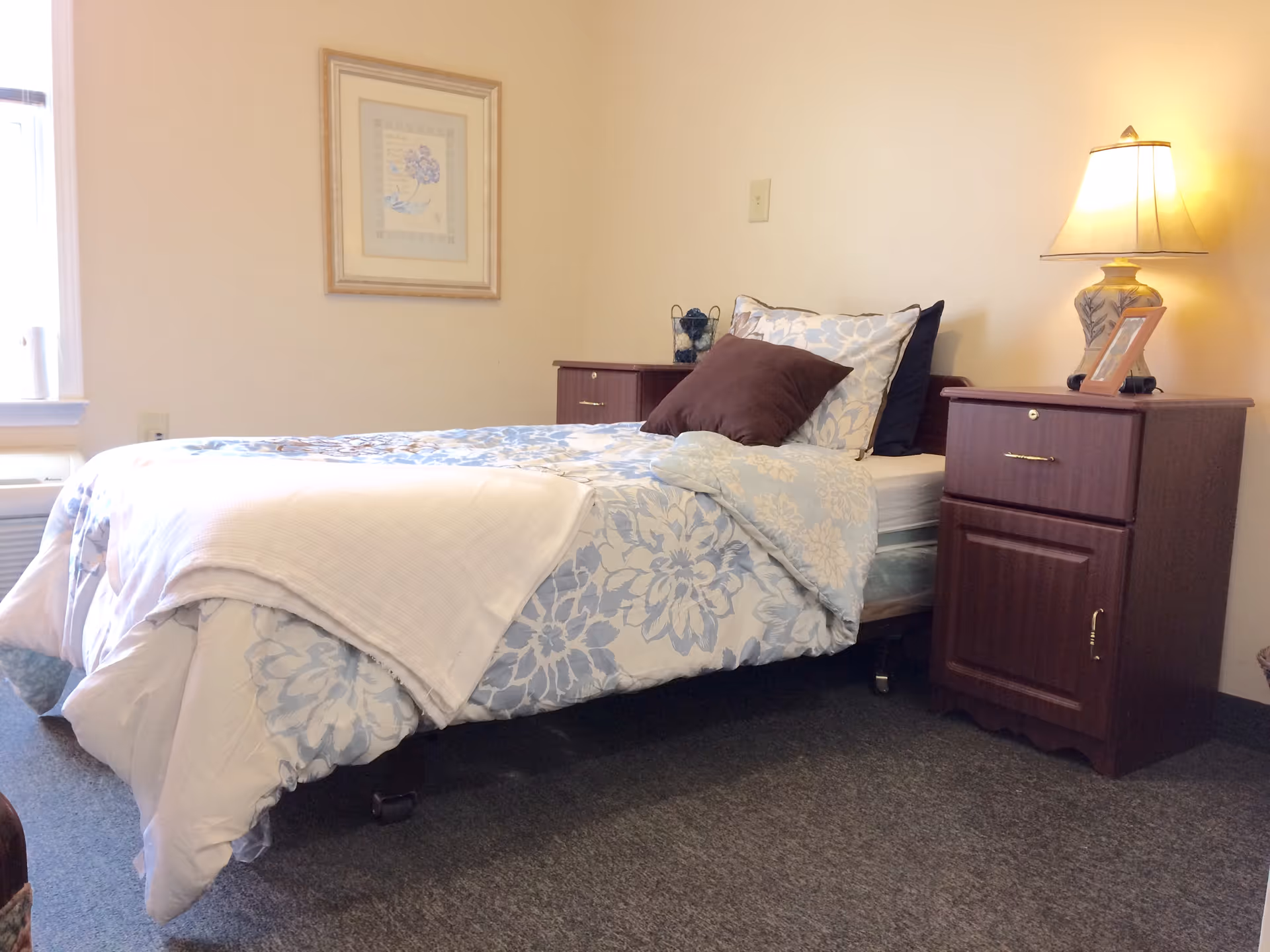 A neatly made single bed with light blue floral bedding, two wooden nightstands and a lamp in a small bedroom.