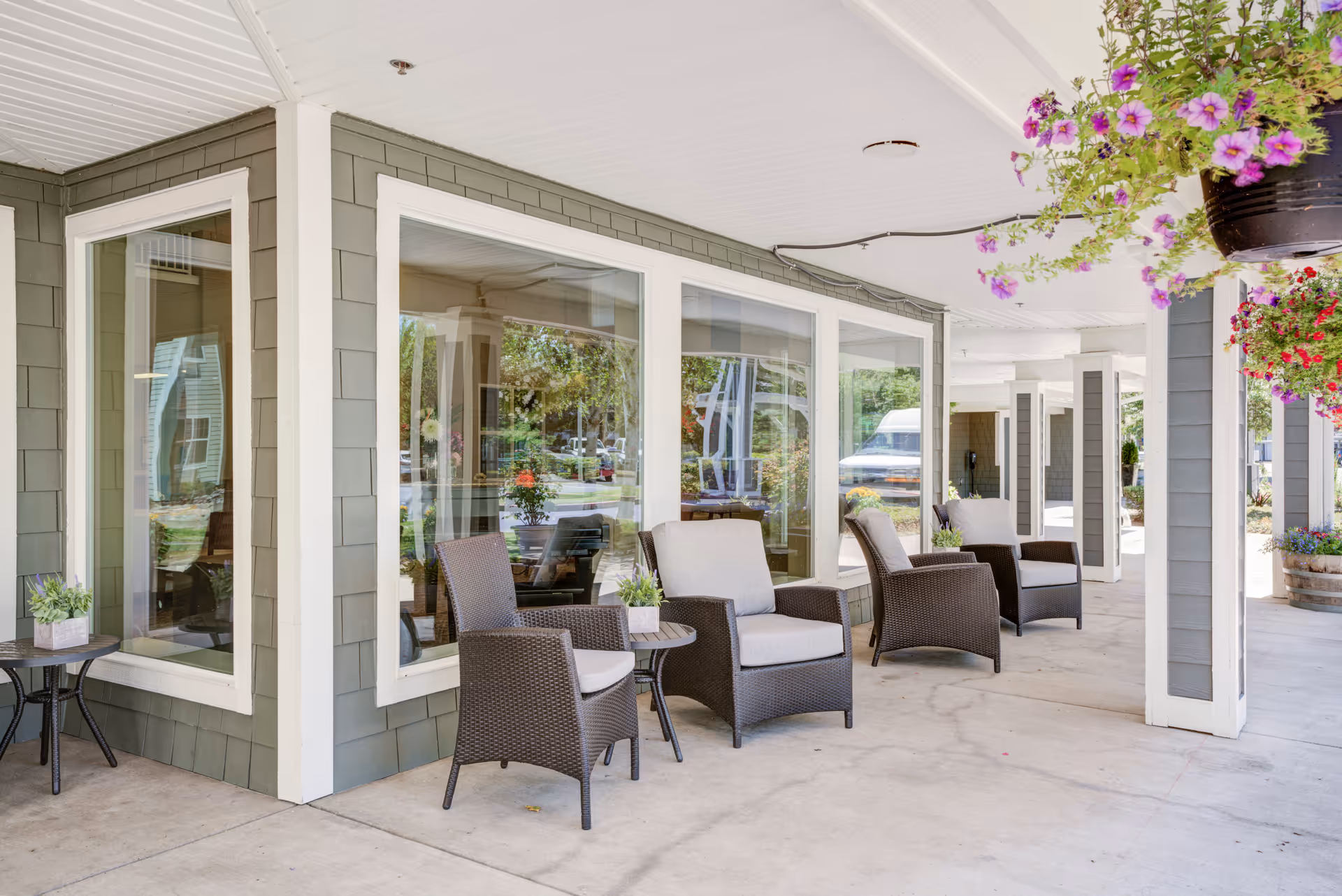 Covered outdoor patio area at Timber Pointe Senior Living with several cushioned wicker chairs and small tables arranged along the building's exterior. Hanging flower baskets with pink and red flowers add color to the space, and large windows provide a view into the interior.