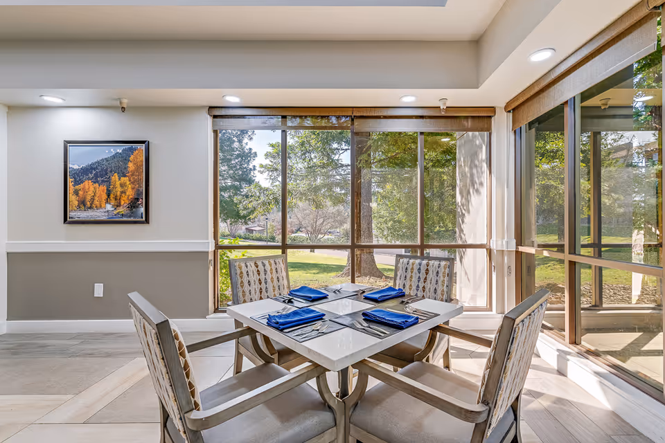 A bright dining area with a square table set for four, featuring blue napkins and silverware. The room has large windows with views of green trees and a lawn outside. A framed picture of a scenic autumn landscape hangs on the wall.