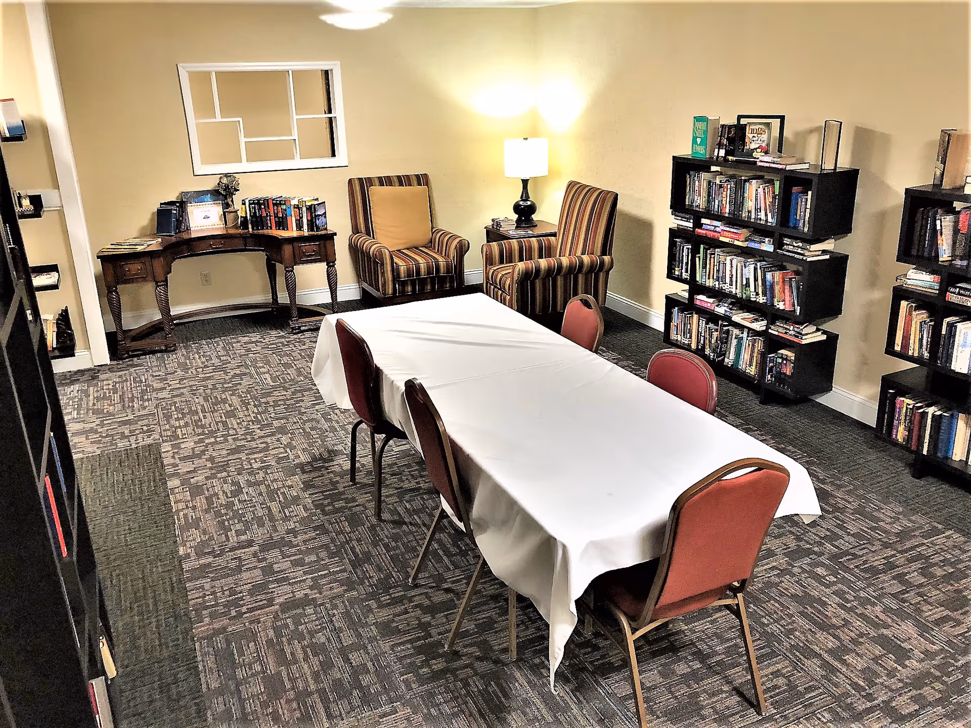 A cozy room with a rectangular table covered with a white tablecloth surrounded by four red chairs. Two striped armchairs with a small side table and lamp are positioned against the wall. Several bookshelves filled with books line the walls, and a wooden desk with books and decor is in the corner. The room has patterned carpet and soft yellow walls.