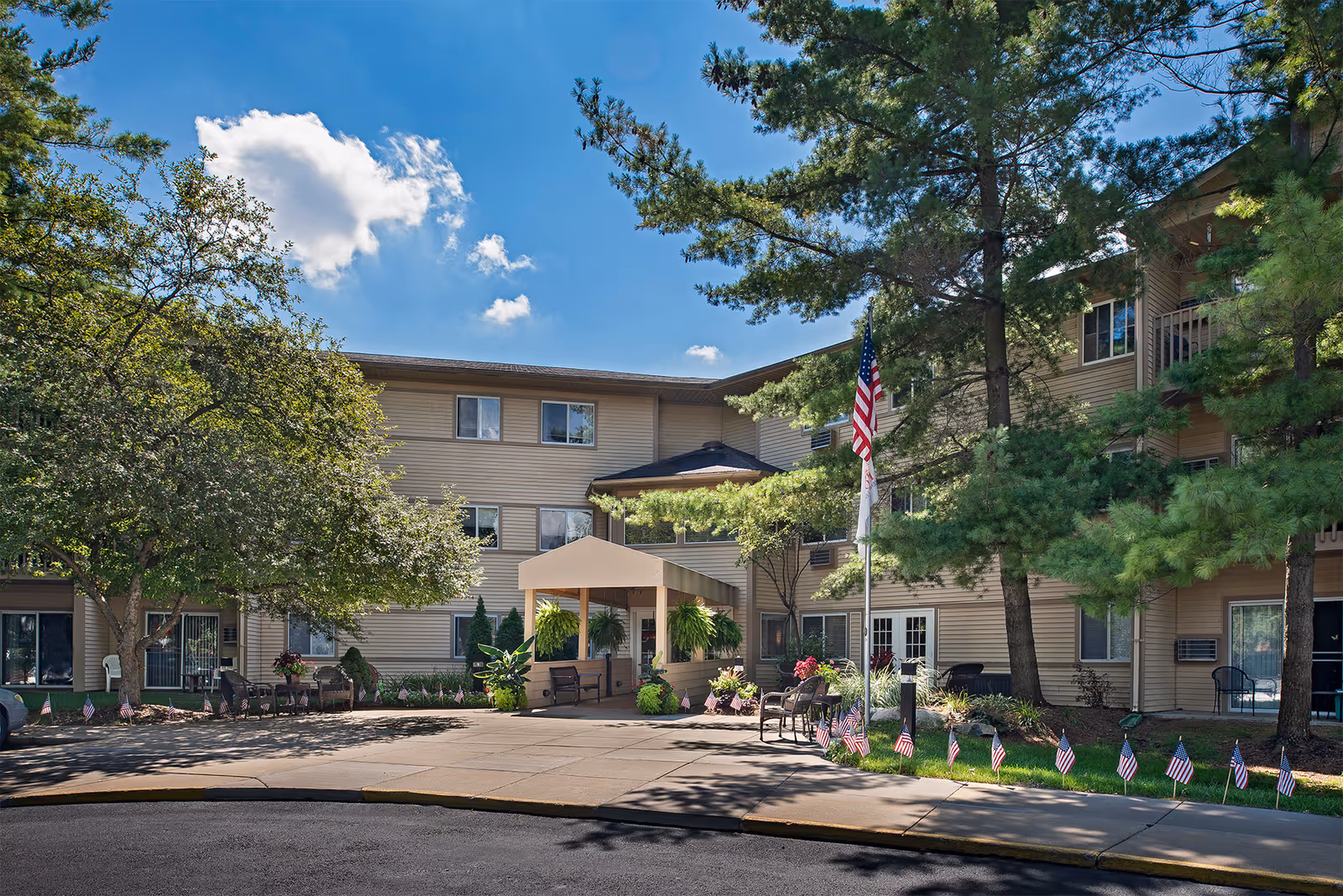Front exterior of a three-story senior living building with a covered entrance, American flag, trees, and small flags lining the walkway.