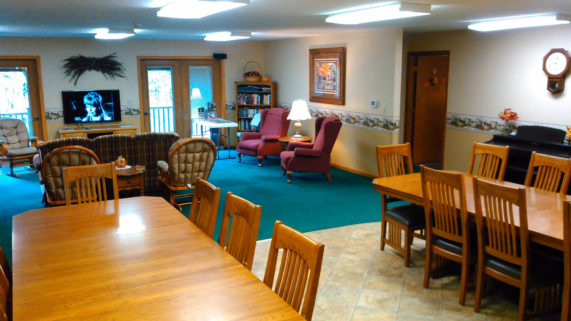 A cozy common area in a senior living facility featuring two wooden dining tables with chairs in the foreground, a green carpeted lounge area with armchairs and a sofa, a television on a stand, a bookshelf, a piano against the wall, and a wall clock. The room has large windows and doors letting in natural light.