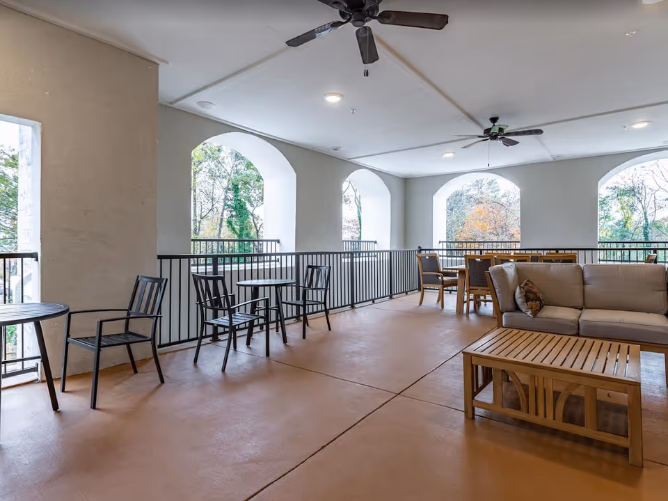 Covered outdoor seating area with tables, chairs, a sofa and ceiling fans overlooking trees through arched openings.