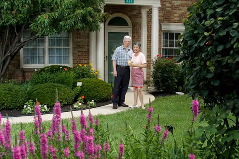 An older couple standing on a curved walkway in front of a brick building entrance surrounded by flowering bushes and a green lawn.