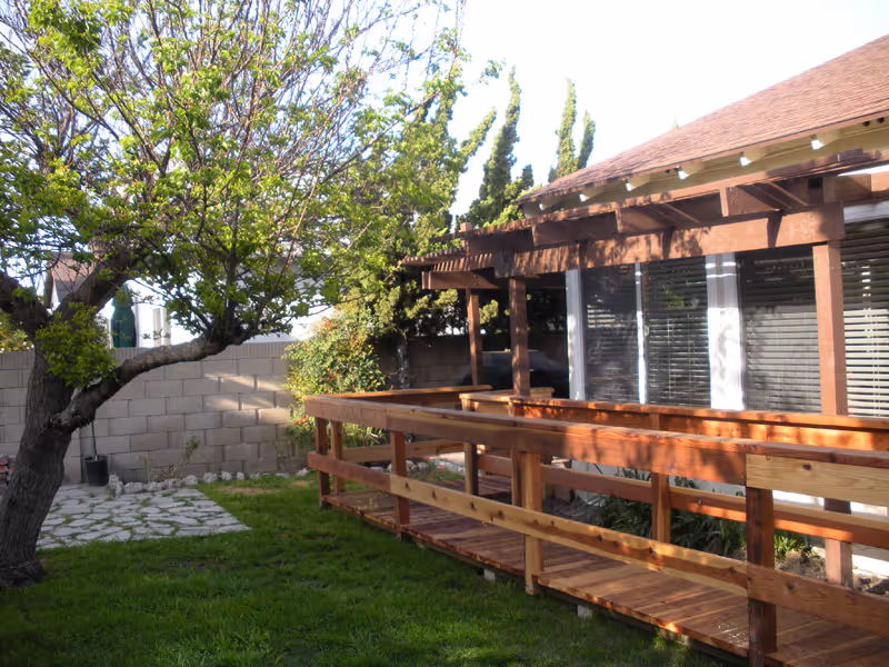 A backyard area with a wooden ramp leading to a house with large windows covered by blinds. There is a tree with green leaves on the left side and a stone pathway near a brick wall in the background.
