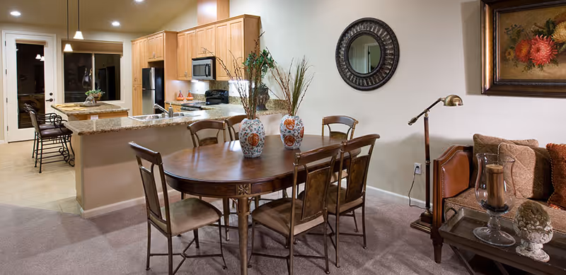 Interior view of a senior living facility showing a dining area with a wooden table and six chairs, decorated with two vases containing dried plants. In the background, there is a kitchen with wooden cabinets, granite countertops, a sink, a microwave, and a refrigerator. To the right, part of a living room is visible with a sofa, a floor lamp, a side table with decorative items, a round mirror on the wall, and a framed painting.