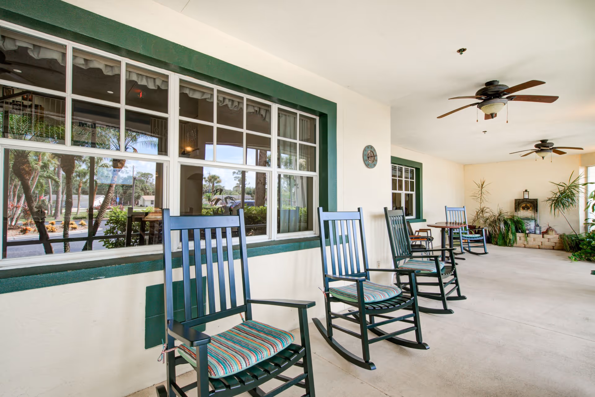 Covered outdoor patio area with several dark green rocking chairs with striped cushions, ceiling fans, potted plants, and large windows reflecting palm trees and outdoor scenery.